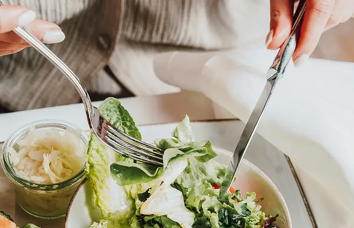Zwei Frauenhände halten Messer und Gabel und machen sich über den Beilagensalat auf dem Tisch her.