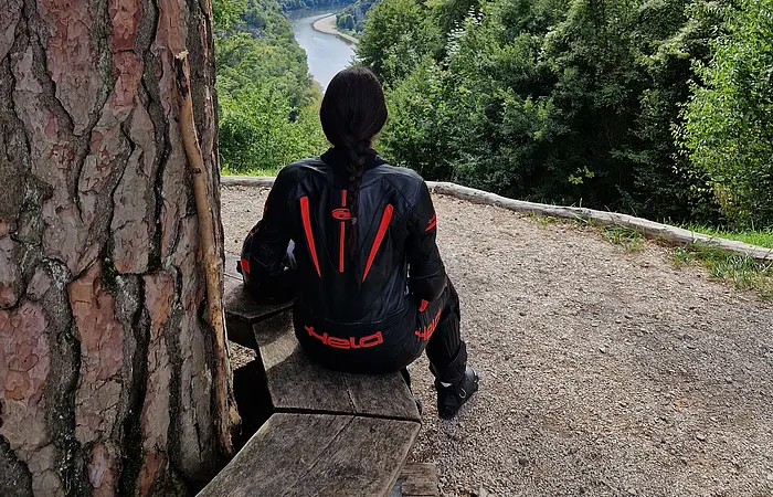 Person mit schwarzer Motorradjacke sitzt auf Holzbänken und blickt auf Fluss und bewaldete Hügel.
