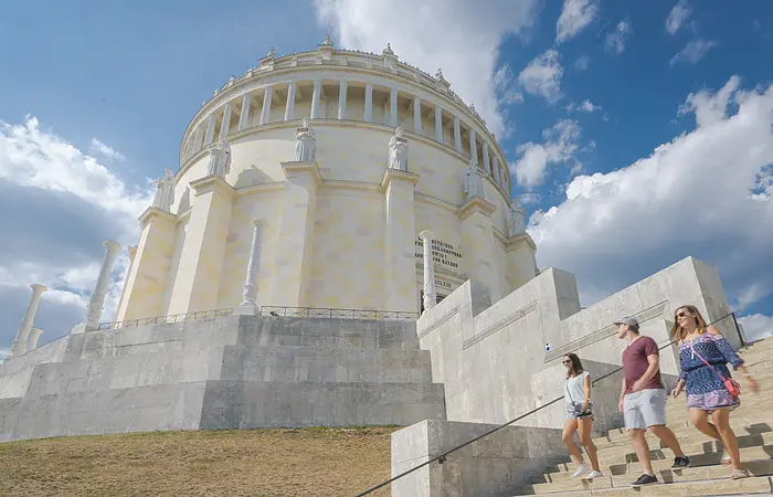 Zwei Frauen und ein Mann, in sommerlicher Kleidung, steigen die Treppen der Befreiungshalle hinunter und werfen einen letzten Blick zu ihr hinauf. Der Himmel strahlt blau und ein paar Wolken sind zu sehen.