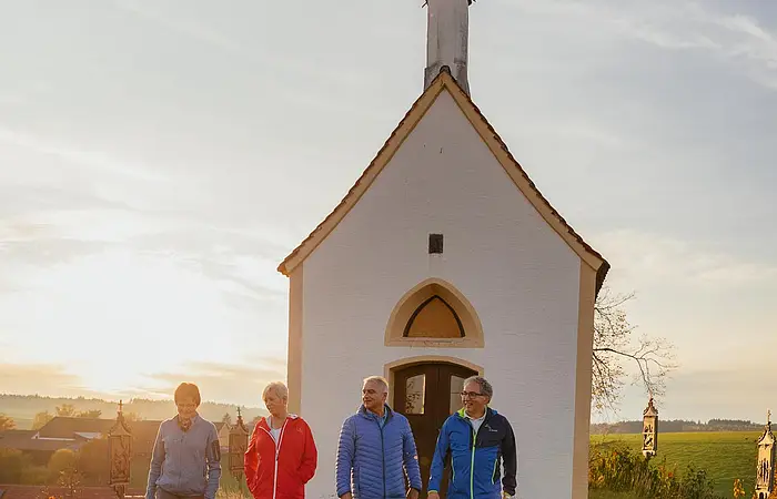 Vier Personen gehen nebeneinander auf einer Wiese vor einer kleinen Kapelle im Sonnenuntergang spazieren. Im Hintergrund grüne Wiese und weitere Gebäude.