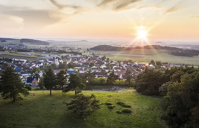 Der Blick von oben über grüne Wiesen hinab auf die Stadt Monheim. Die Sonne strahlt am Horizont und ist fast untergegangen.