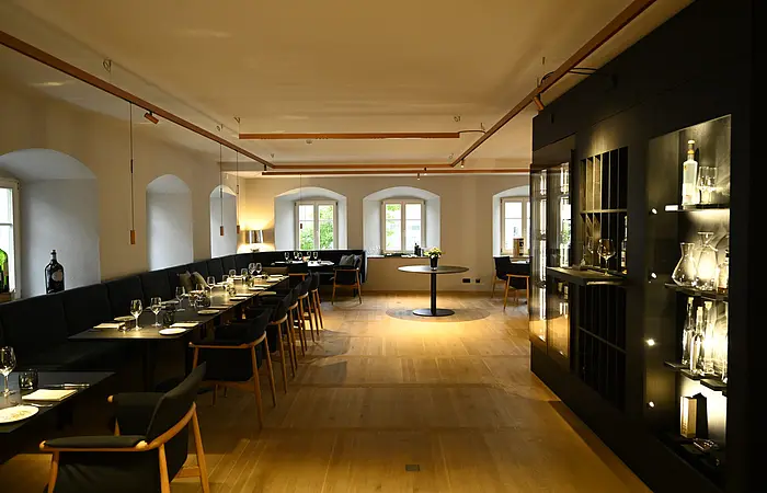 Innenansicht des Restaurants im Hotel by Ferber, in Monheim. Auf der rechten Seite ein großes schwarzes Regal mit verschiedenen Glasbehältern, Weingläsern und Flaschen. Auf der linken Seite schwarze Holztische mit schwarzen Sitzbänken und schwarzen Stühlen zu sehen.