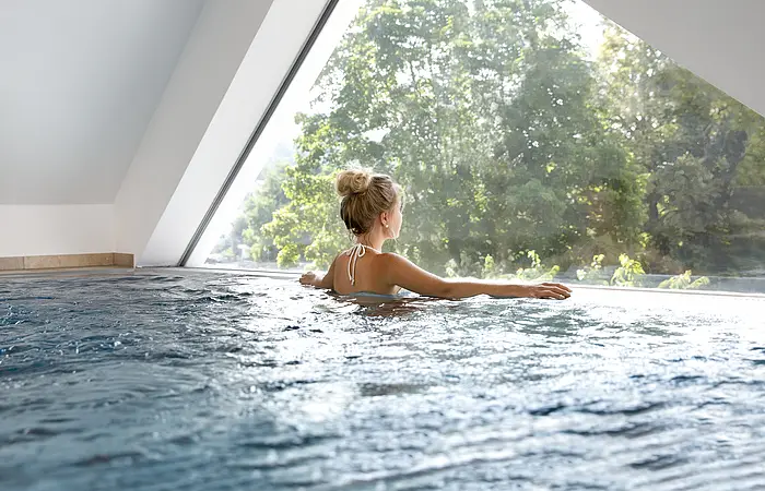 Ein Gast genießt den Ausblick von Schwimmbecken durch ein Fenster nach draußen in die Naturlandschaft.