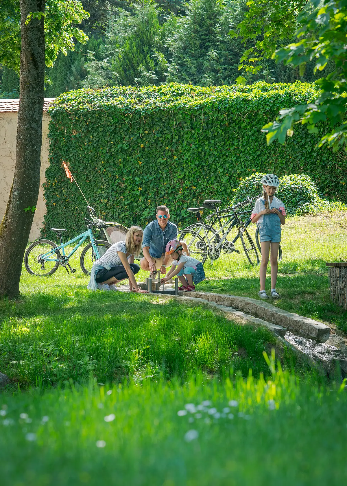 Eine Familie mit Fahrrädern spielt im Park neben einem kleinen Wasserlauf.
