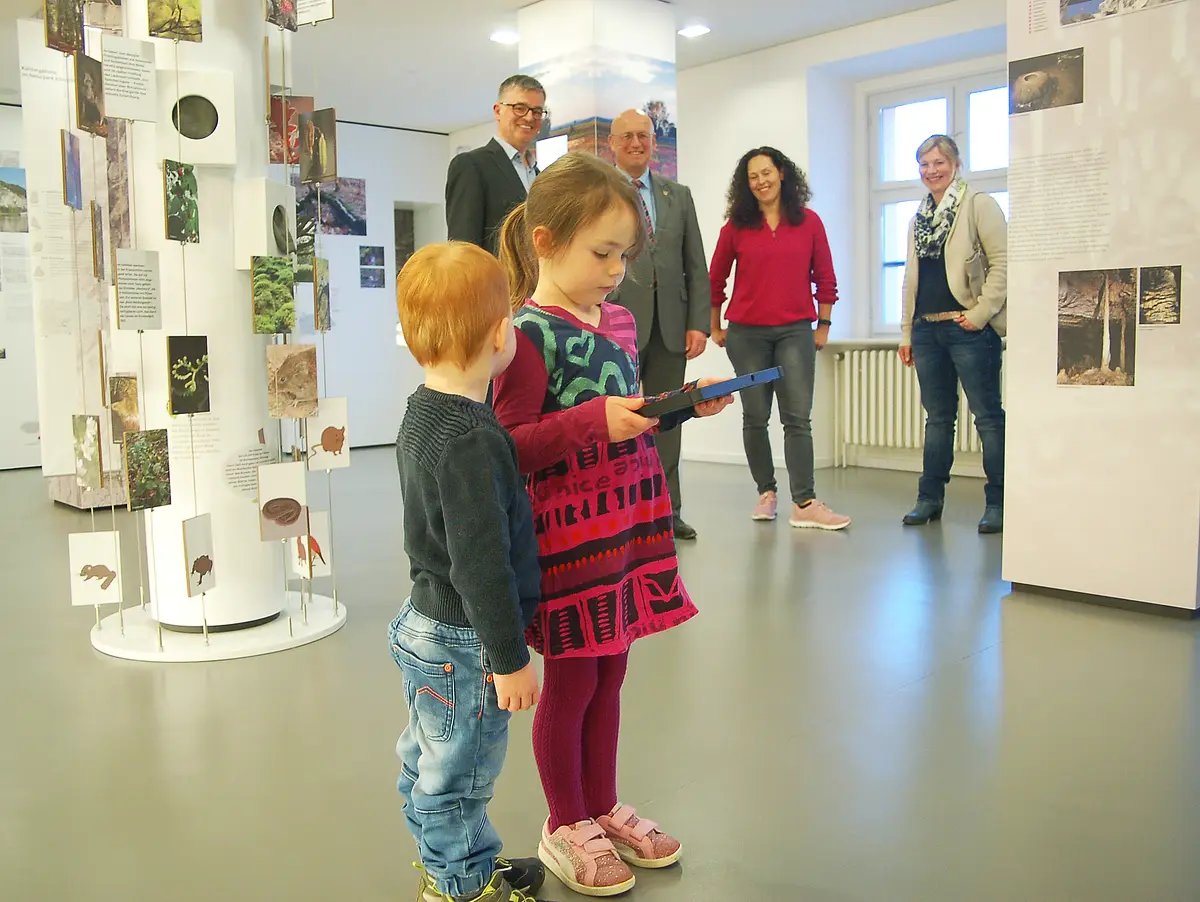Ein Junge und ein Mädchen laufen mit einem Tablet durch die Ausstellung im Informationszentrum. Das Tablet spielt den Audiguide vor. Eine lustige Schnitzeljagt mit Fossi und Juvi durch das Museum. Im Hintergrund stehen Christoph Würflein und Anton Knapp.