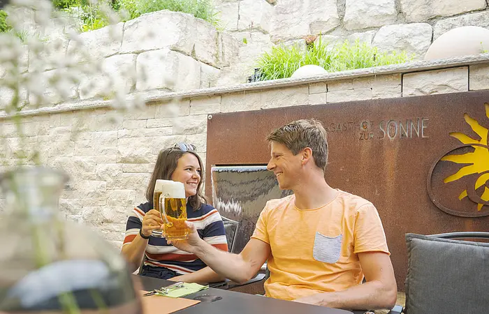 Zwei Personen stoßen mit Biergläsern an, sitzend vor einer Steinmauer mit Schriftzug „Gasthof zur Sonne“.