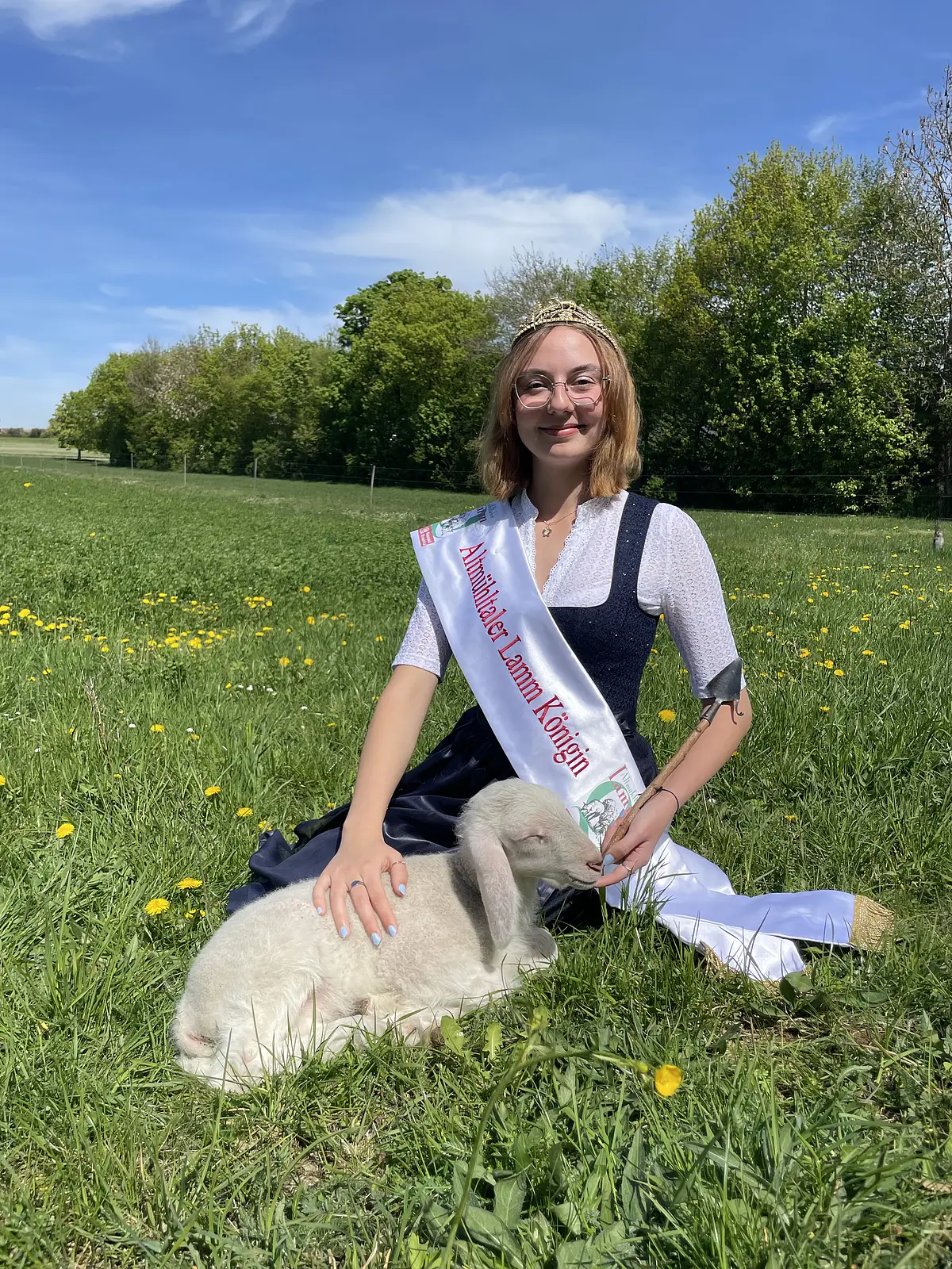 Altmühltaler Lammkönigin Anna Wiest Junge Frau mit Krone und Schärpe sitzt auf Wiese und hält ein weißes Lamm im Arm.