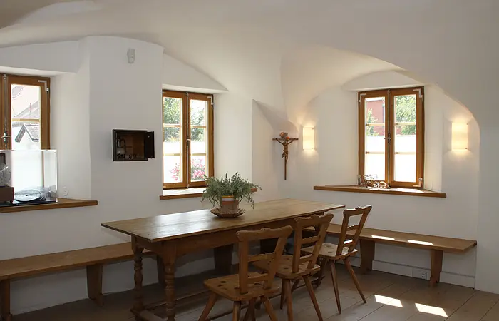 Esszimmer mit Holztisch, Stühlen und Bänken. Zwei Fenster, weiße Wände, Pflanze auf dem Tisch.