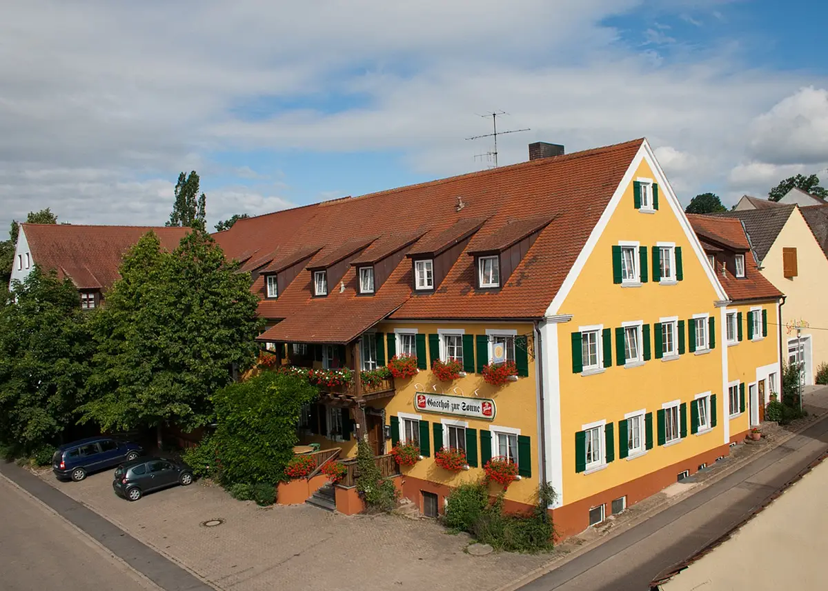 Ein großes langes vierstöckiges Haus im fränksichen Baustil mit gelber Fassade und grünen Fensterläden. An den Fenster an der Front hängen Kästen mit roten Blumen.