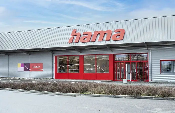 Graue Fassade eines Outlet-Geschäfts mit rotem Schriftzug "Hama", roten Eingangstüren und roten Fensterumrandungen, bei bewölktem Himmel. Vor dem Geschäft eine Hecke entlang einer Straße.
