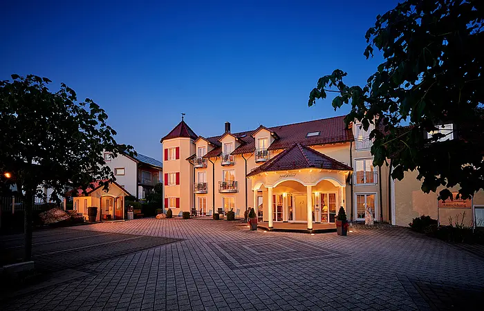 Beleuchtetes Hotelgebäude mit gepflastertem Vorplatz und Bäumen bei Abenddämmerung.