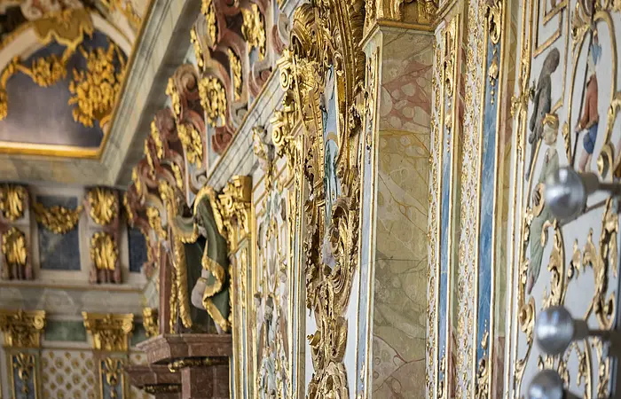 Detailaufnahme einer Wand mit barocker goldener Stuckverzierung und bemalten Figuren in einem historischen Gebäude.