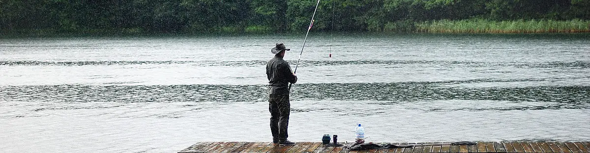 Person steht auf Holzsteg und angelt an einem See mit bewaldetem Ufer im Hintergrund.