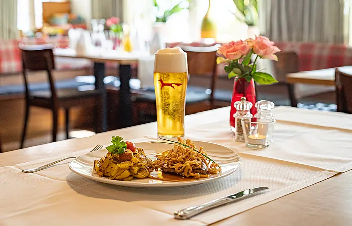 Am Tisch mit beigem Läufer steht ein Helles und auf dem Teller davor ist ein Zwiebelrostbraten mit Bratkartoffeln angerichtet. Im Hintergrund sind weitere Tische zu erahnen.