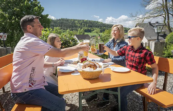 Biergarten, Nahaufnahme einer Familie mit zwei jüngeren Kindernv prosten sich bei einer Brotzeit mit Brezen, Käse und Radieschen zu.