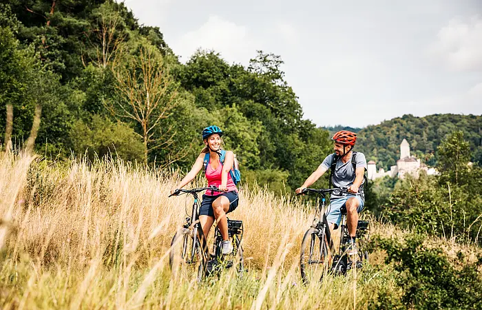 Zwei Radler, eine Frau und ein Mann, sind auf dem Radweg unterwegs. Die Gräser entlang des Radweges sind recht schon recht vertrocknet und braun. Dahinter blitzt in weiß die Burg Kipfenberg inmitten des Waldes heraus.