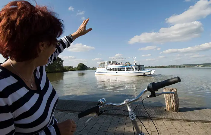 Eine Fahrradfahrerin winkt vom Ufer aus der MS Altmühlsee zu.