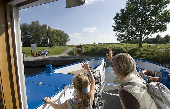 Eine Frau und ein junges Mädchen winken vom Schiff den Besuchern am grünen Uferrand.