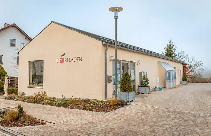 Eingang eines hellen Gebäudes mit Schriftzug „Dorfladen“ und Pflanzkübeln vor der Tür.