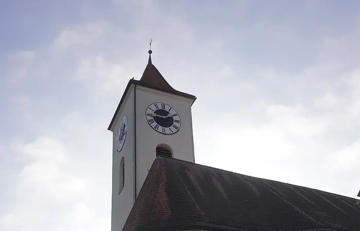 Kirchturm mit Uhr und weißem Gebäude bei bewölktem Himmel in einem Dorf.