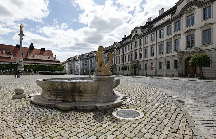 Die Aufnahme zeigt den Residenzplatz, wobei sich der Blick rechts auf eine lange Reihe von mehrstöckigen Häuserfassaden richtet.
Der Residenzplatz selbst ist durchgehend mit Kopfsteinpflaster bedeckt. Im Hintergrund befindet sich ein Brunnen, in dessen Mitte auf einer hohen Säule eine vergoldete Marienstatue thront. Links davon ist die Residenz im Blickfeld, die allerdings nicht vollständig im Bild ist. Unmittelbar vor dem Betrachter steht auf der rechten Bildseite ein zweiter kleiner Brunnen.
