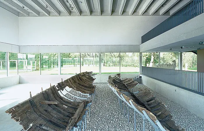 Zwei antike Holzbootwracks auf Kiesboden in einem modernen Ausstellungsraum mit Glaswänden.