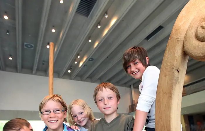 Fünf Kinder stehen in einem hölzernen Boot in einem Gebäude mit Betonwänden und Deckenbeleuchtung.