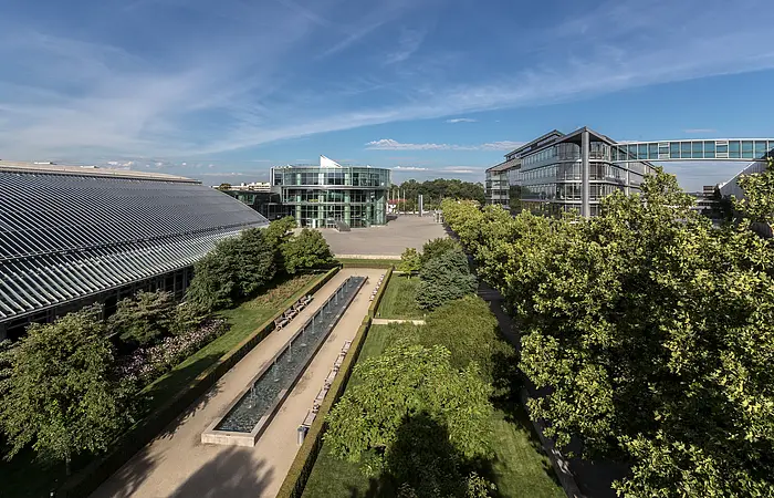 Das AUDI Forum von weitem und etwas erhöht liegt es idyllisch umgeben von Bäumen und Sträuchern. Der Himmel erstrahlt blau.
