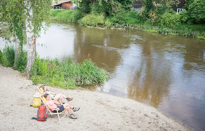 Zwei Personen mit Rucksäcken neben sich  sitzen auf Liegestühlen am Flussufer neben Bäumen an einem sonnigen Tag.
