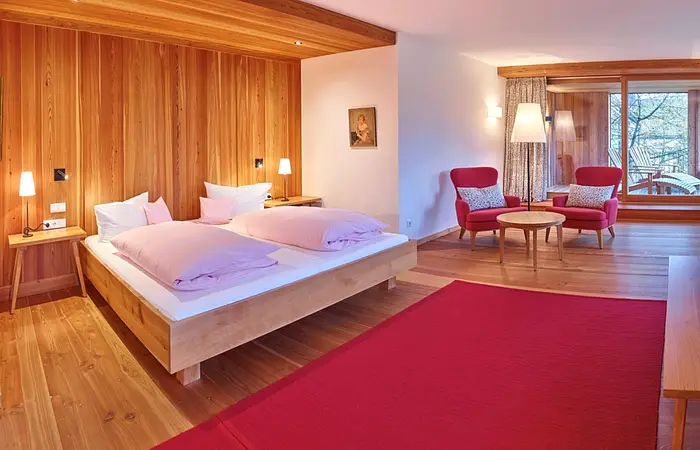 Schlafzimmer mit Holzverkleidung, Doppelbett, zwei roten Sesseln, Tisch, Stehlampe und großem Fenster mit Blick auf Terrasse.
