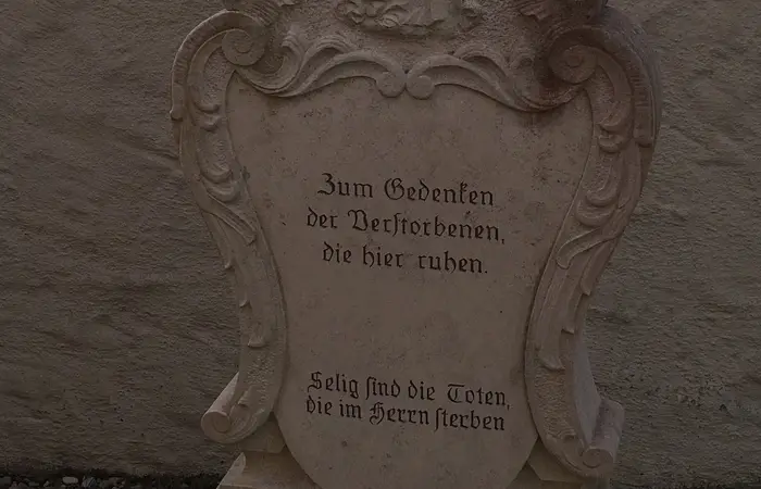 Grabstein mit Relief einer stehenden Figur und der Inschrift „Zum Gedenken der Verstorbenen, die hier ruhen“ in deutscher Sprache.