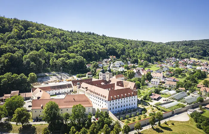 Luftaufnahme eines großen Gebäudekomplexes mit roten Dächern vor bewaldeten Hügeln und einem Dorf im Hintergrund.