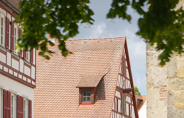 Fachwerkhaus mit rotem Dach und weißen Wänden, umgeben von Bäumen. Eine Familie geht auf einem gepflasterten Weg vorbei.