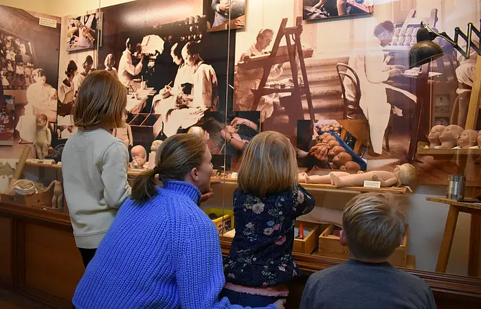 Eine Frau sieht sich mit drei Kindern eine Vitrine im Käthe-Kruse-Puppenmuseum an in der erklärt wird, wie die Puppen hergestellt werden.