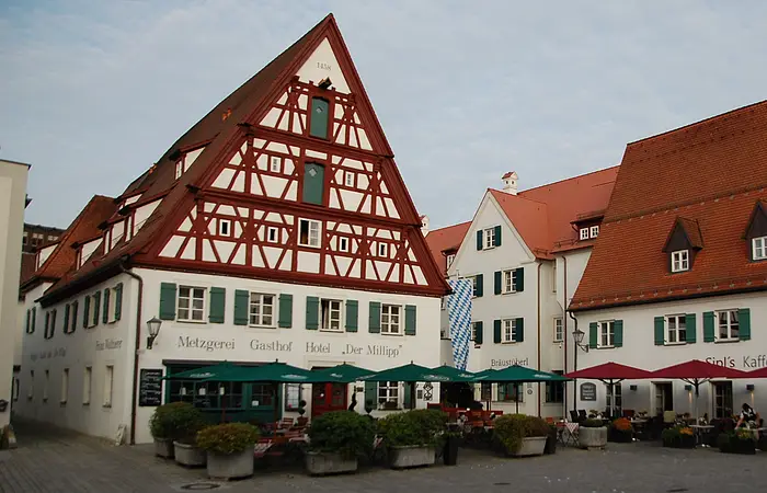 Traditionelle Fachwerkhäuser mit Außenbestuhlung und Sonnenschirmen an einem gepflasterten Platz.