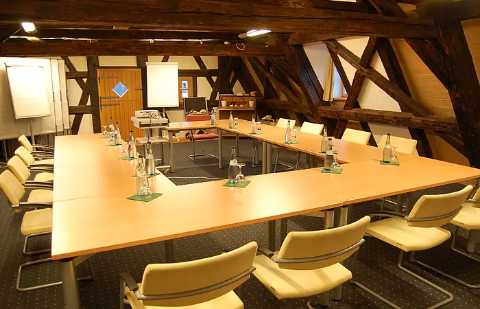 Konferenzraum mit U-förmigen Tischen, Stühlen, Wasserflaschen und Flipcharts unter Holzbalken.
