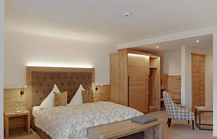 Hotelzimmer mit Doppelbett, Holzmöbeln, Sessel und Tisch in neutralen Farben und Beleuchtung.