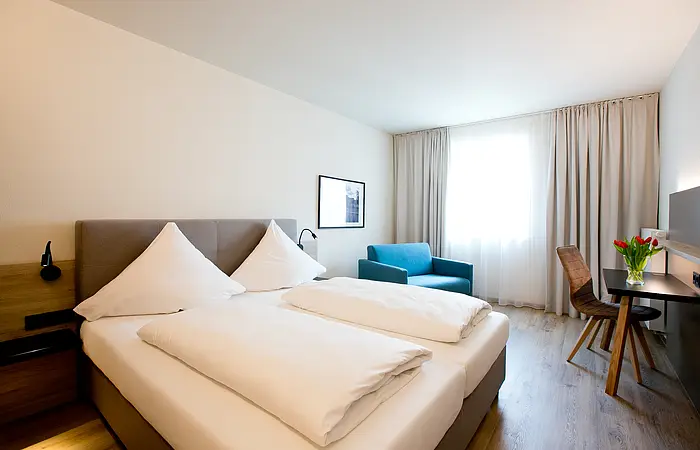 Hotelzimmer mit zwei Einzelbetten, blauem Sessel, Schreibtisch mit Stuhl und Fenster mit Vorhängen.