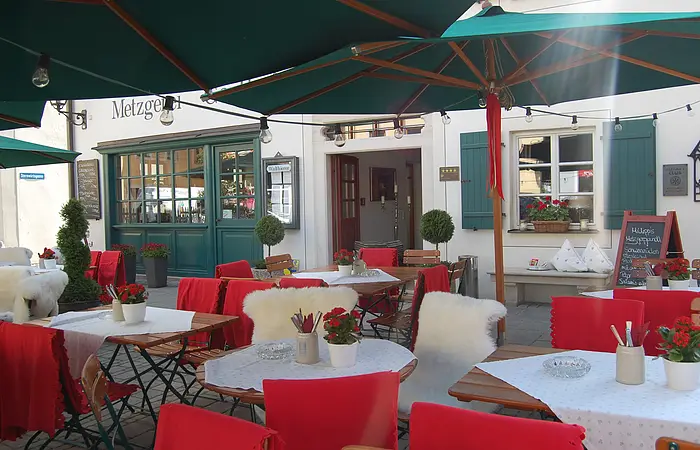 Außenbereich eines Cafés mit roten Stuhlhussen, weißen Tischdecken und grünen Sonnenschirmen.