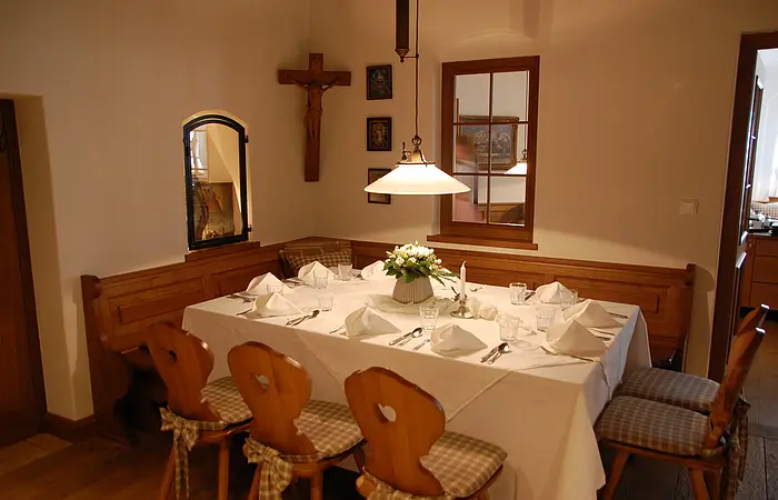 Esszimmer mit Holztisch, weißer Tischdecke, sechs Holzstühlen mit Kissen, Wandkreuz und Fenster mit Spiegel.