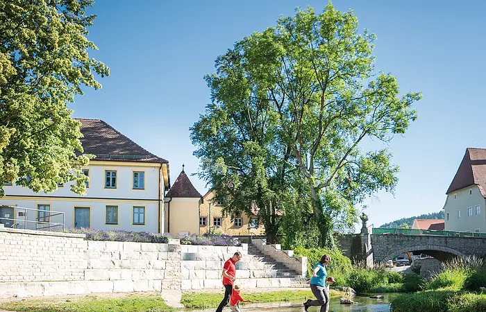 Zwei Personen überqueren auf Trittsteinen einen flachen Fluss, im Hintergrund Gebäude und Bäume.