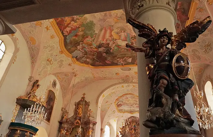 Innenraum einer Kirche mit bemaltem Gewölbe und Statue eines Engels mit Schild und Schwert an einer Säule.