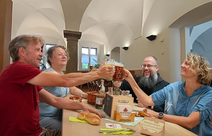 Vier Personen, zwei Frauen und zwei Männer stehen um eine Holztheke, und prosten sich mit einem Krug Bier zu. Auf der Theke stehen eine Kiste mit Besteck und ein kleiner Korb. Es liegen zwei Semmeln und eine Breze daneben. Im Hintergrund sieht man Säulen und Bügen des Klostegewölbes.