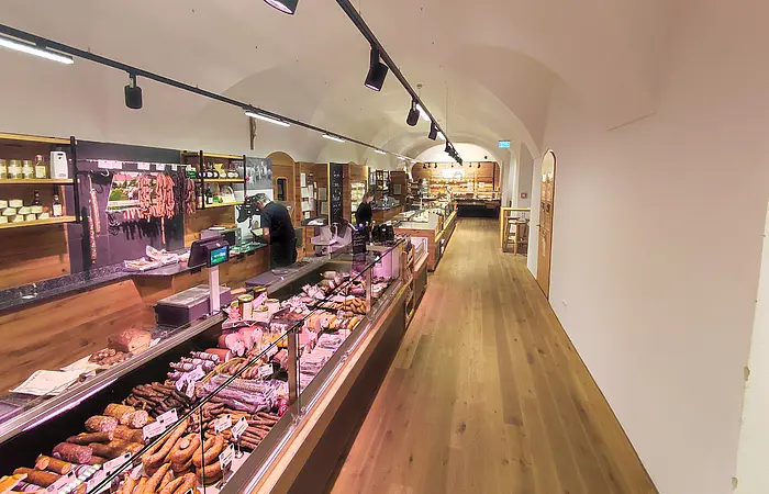 Eine Fleisch- und Wursttheke mit Inhalt seitlich aufgenommen. Dahinter ein Holzregal mit verschiedenen Waren.