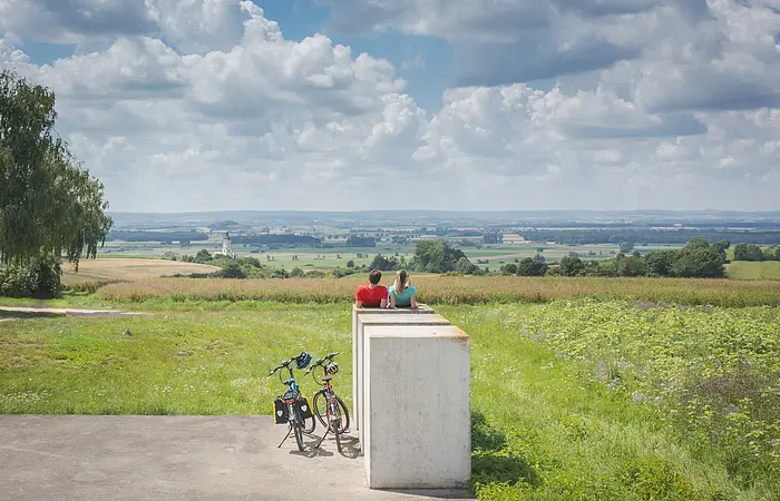 Zwei Personen sitzen auf einer Betonbank und blicken auf eine weite Landschaft mit Feldern und einem Kirchturm. Zwei Fahrräder stehen daneben.