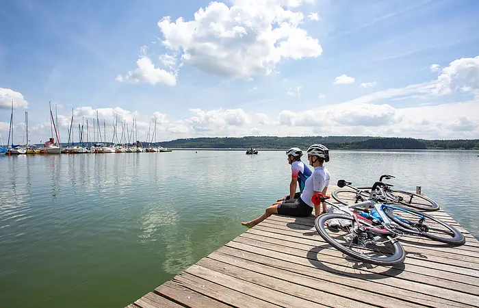 Zwei Rennradler machen Pause am Brombachsee. Sie sitzen dabei auf einem Holzsteg und blicken zu den Segelbooten