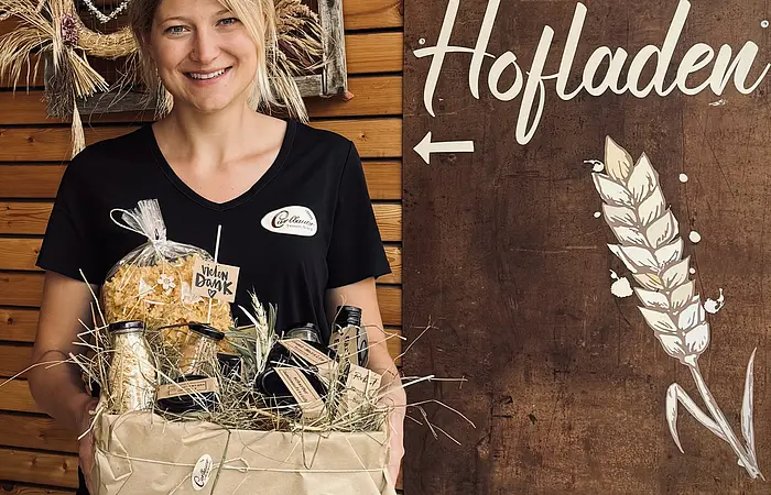 Frau mit Geschenkbox, in der regionale Produkte sind, vor Holzhaus und Schild mit Wegweisern zu Fahrradgarage und Hofladen