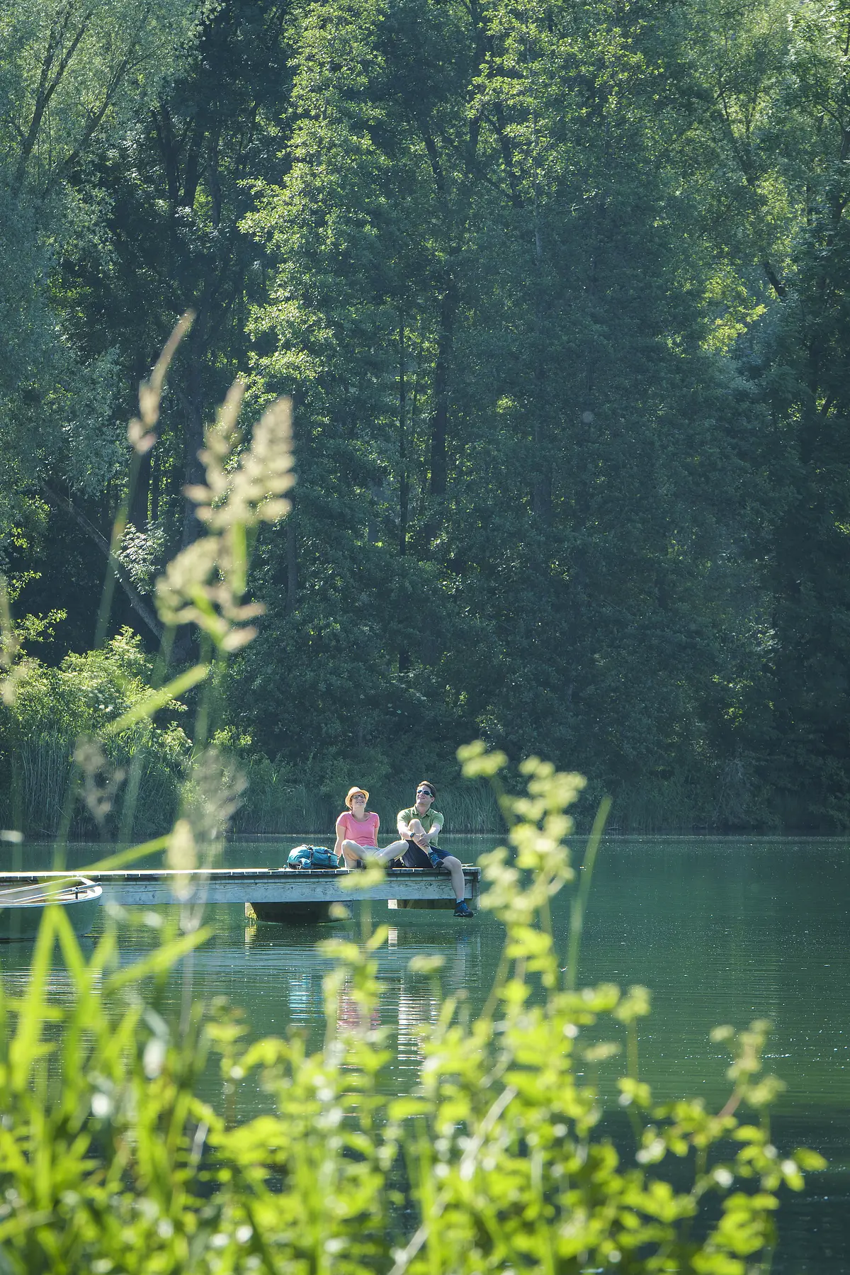 Ein Wanderpärchen sitzt auf dem Steg am Kratzmühlsee und lässt den Blick über den See schweifen. Hinter ihnen ragen die Bäume mit ihrem grünen Blätterkleid auf und links am Steg ist ein Ruderboot befestigt.