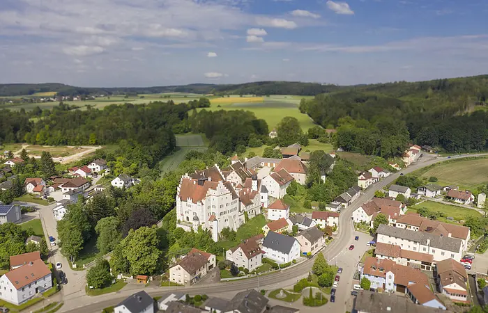 Luftaufnahme eines Dorfes mit einem großen weißen Gebäude im Zentrum, umgeben von Bäumen und Feldern.