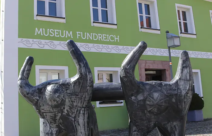 Außenansicht des Archäologischen Museums in Thalmässing. Grünes Haus mit schwarzem Dach. Davor steht eine Skulptur von menschlichen Figuren, die eine Platte tragen.